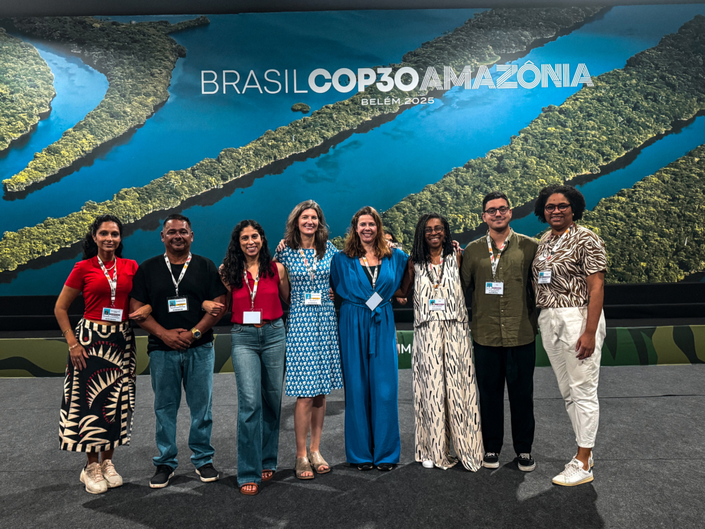A group of colourfully dressed people from Amnesty International stand against a backdrop of waterways. The sign reads Brasil COP30 Amazonia.