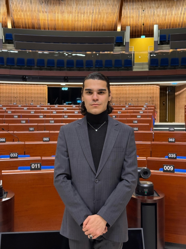 A person with slicked back hair, wearing a grey suit and black poloneck, looks to camera