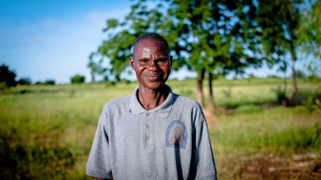 A smiling man, stands in a sunfilled field.