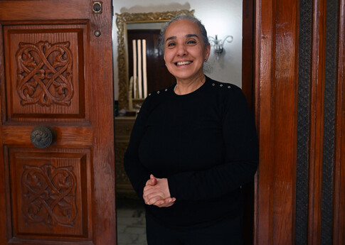 A smiling lady stands between two wooden doors, hands clasped. 