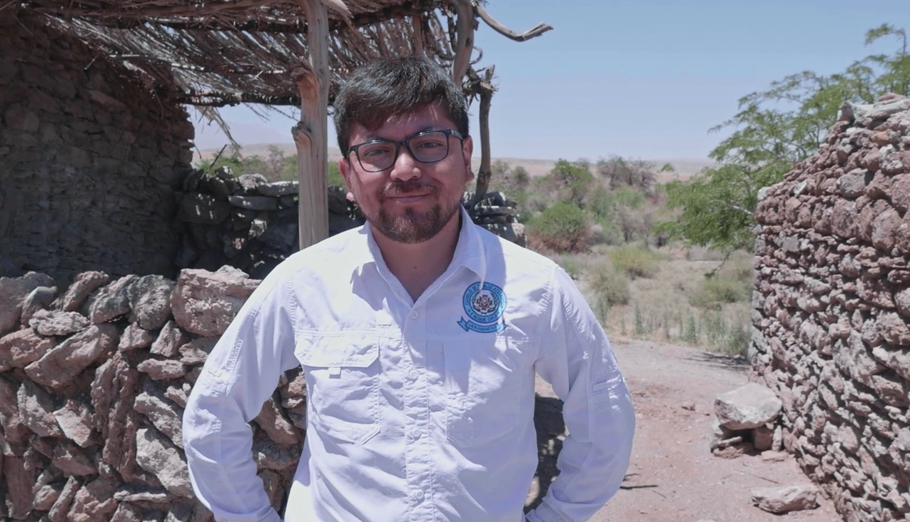 A man in a white shirt and glasses stands in front of a stone wall