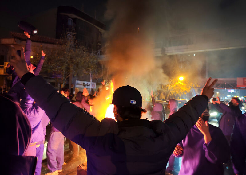 A protester stands with both arms in the air, silhouetted against a fire burning in the background.