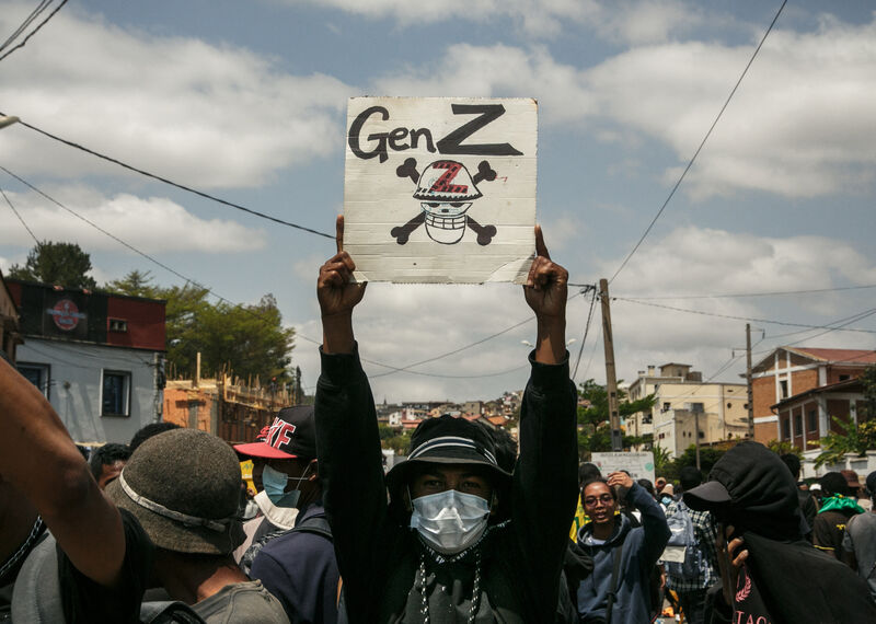 Amongst a crowd of people, a protester, wearing a hat and face covering, holds up a placard with GenZ and skull and cross bones. 