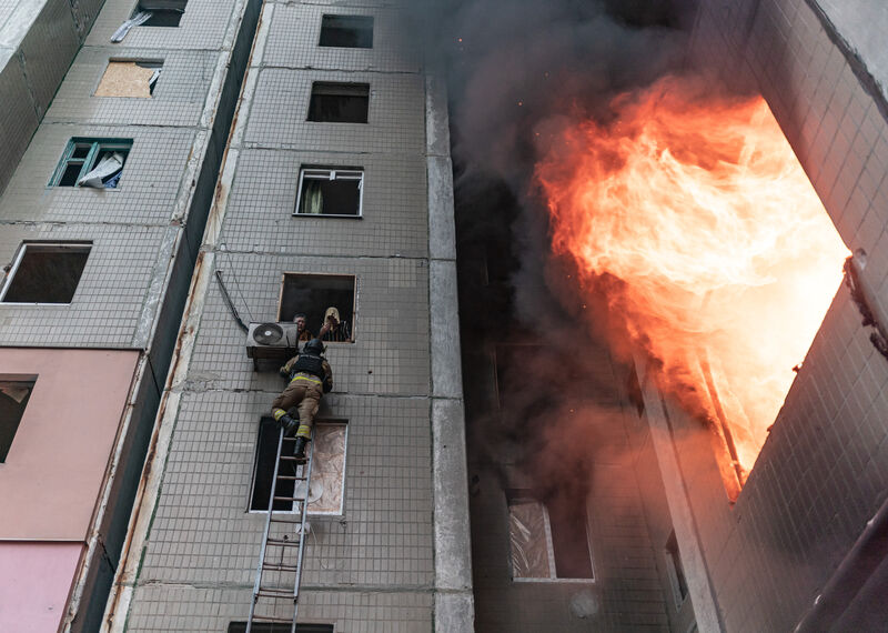 Looking up at a tall building with a large fire raging as firefighter climbs up a ladder towards two people leaning out of one of the windows.