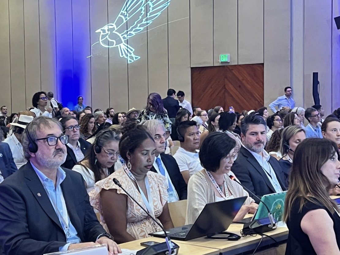 A large group of people sit in rows at a conference. Some wear headsets, take notes, or use laptops. A glowing bird emblem is projected on the wall behind them. The atmosphere is attentive and formal.