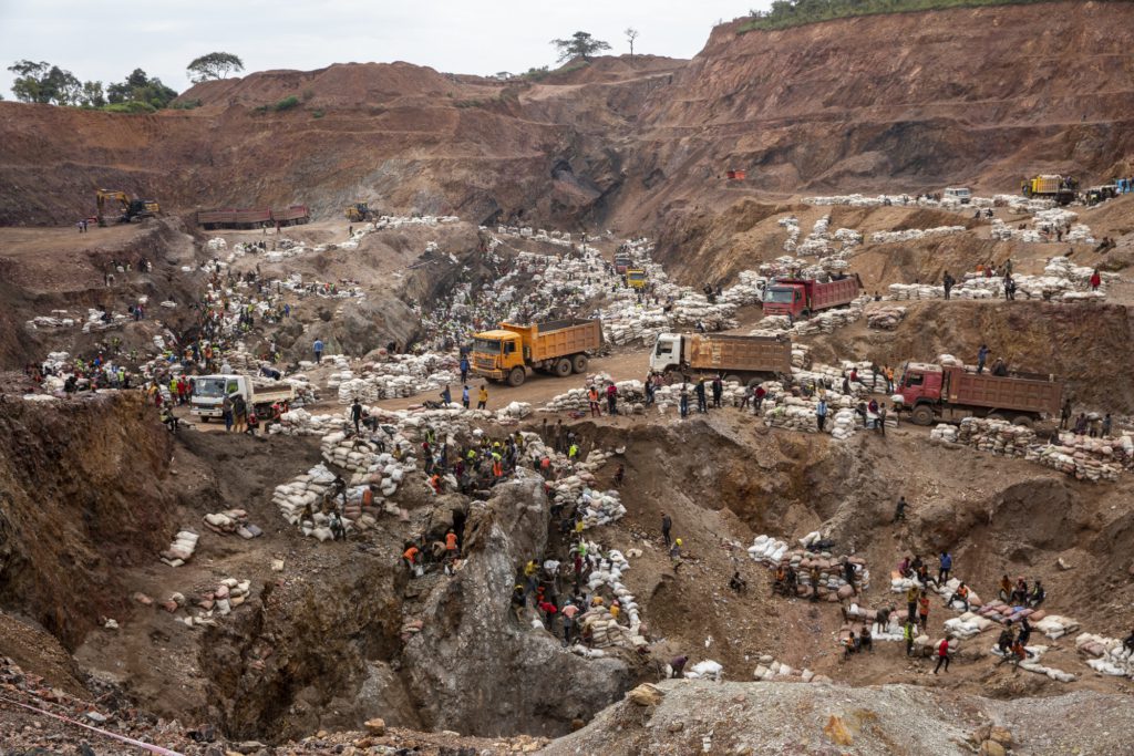 a drone camera image of a cobalt mine in the DRC. The area is surronded by trucks and hundreds of workers and stacks of bags.
