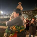 A man in a grey jumper warmly embraces an older woman outside an official looking building.