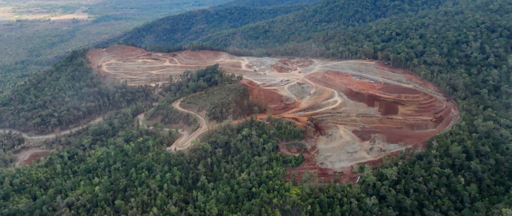 A long-range aerial shot of a forested area on a hillside. At the centre of the image is a large expanse of red, orange and brown earth, where an arc of trees has been removed to make way for a nickel mine. On the ground you can just make out vehicles and heavy machinery on the site